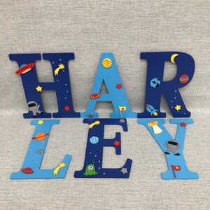 May include: Blue and light blue wooden letters spelling out the name "HARLEY" decorated with space-themed embellishments, including stars, planets, rockets, and aliens.