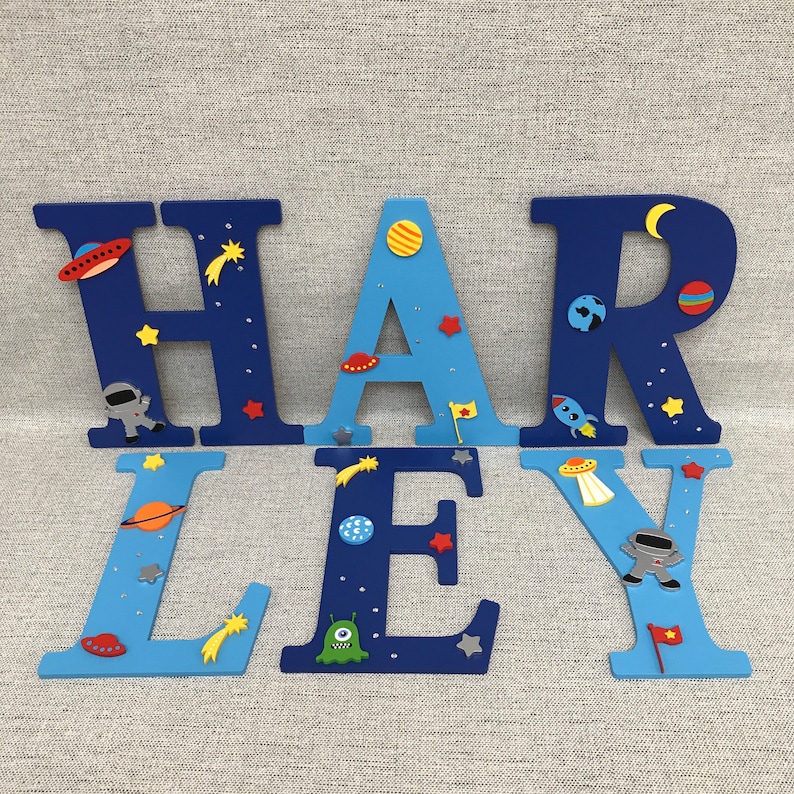 May include: Blue and light blue wooden letters spelling out the name "HARLEY" decorated with space-themed embellishments, including stars, planets, rockets, and aliens.