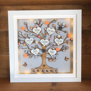 May include: A white framed illuminated family tree with a gray background. The tree is decorated with peach and gray flowers, pearls, and glitter. The names Brian, Caroline, Danielle, James, Ben, Sam, and Nicole are written on the hearts. The words "G₂ R, A, N, NY 4" are spelled out with wooden letter tiles at the bottom of the frame.