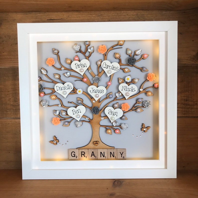 May include: A white framed illuminated family tree with a gray background. The tree is decorated with peach and gray flowers, pearls, and glitter. The names Brian, Caroline, Danielle, James, Ben, Sam, and Nicole are written on the hearts. The words "G₂ R, A, N, NY 4" are spelled out with wooden letter tiles at the bottom of the frame.