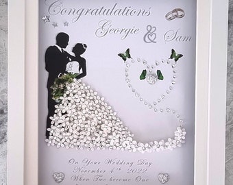 Könnte beinhalten: Ein gerahmtes Hochzeitskunstwerk mit weißem Rahmen. Das Kunstwerk zeigt eine Silhouette von Braut und Bräutigam, ein Kleid mit weißen Blumen und die Worte "Congratulations Georgie & Sam". Verziert mit Schmetterlingen und herzförmigen Verzierungen.