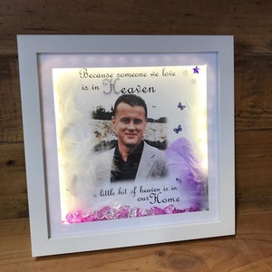 May include: A white framed picture with a photo of a man, surrounded by white and purple feathers. The picture has a light inside and pink and clear crystals at the bottom. The text on the picture reads "Because someone we love is in Heaven, a little bit of heaven is in our Home."