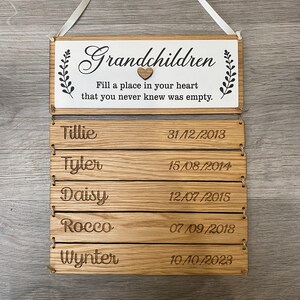 May include: Wooden sign with the text "Grandchildren Fill a place in your heart that you never knew was empty."  Below the text are five lines with names and dates: Tillie 31/12/2013, Tyler 15/08/2014, Daisy 12/07/2015, Rocco 07/09/2018, Wynter 10/10/2023.