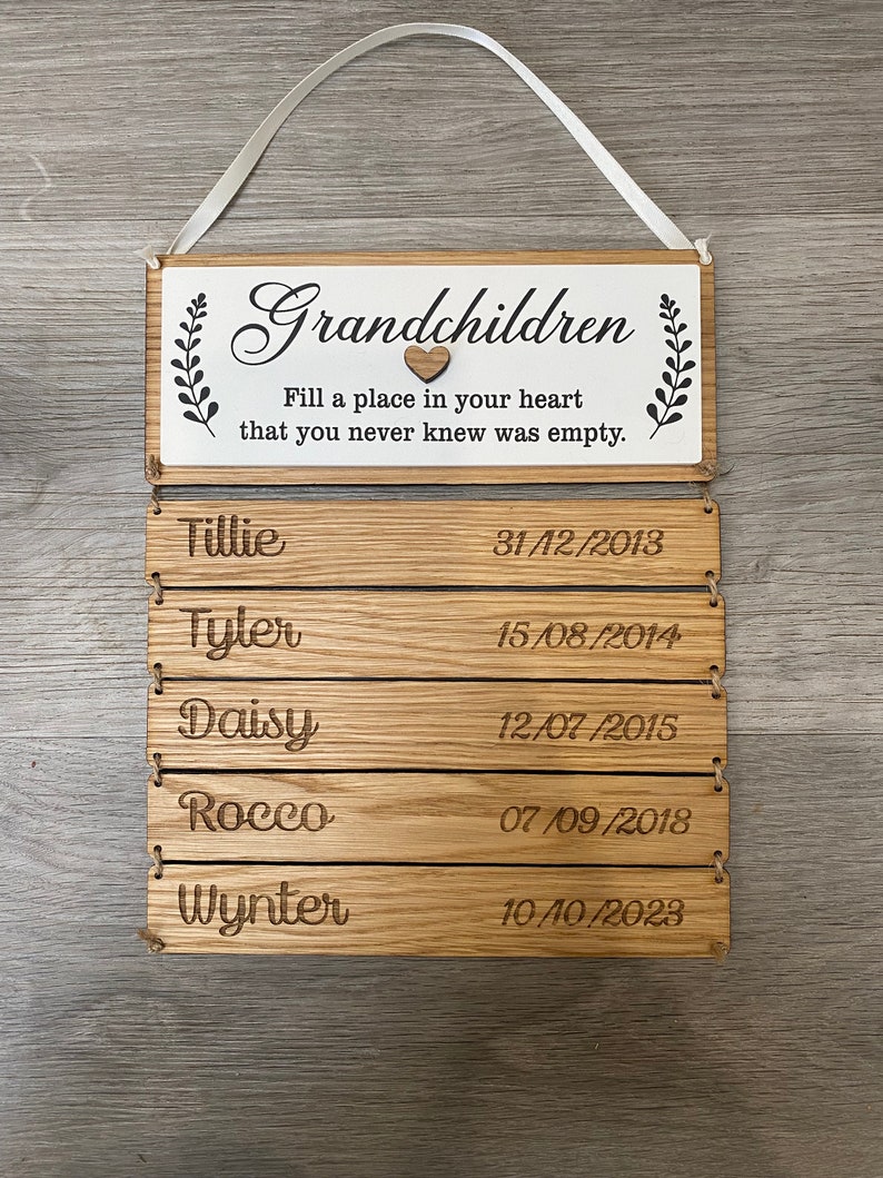 May include: Wooden sign with the text "Grandchildren Fill a place in your heart that you never knew was empty."  Below the text are five lines with names and dates: Tillie 31/12/2013, Tyler 15/08/2014, Daisy 12/07/2015, Rocco 07/09/2018, Wynter 10/10/2023.