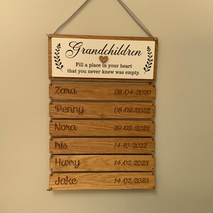 May include: Wooden sign with the text "Grandchildren Fill a place in your heart that you never knew was empty."  The sign has a small heart cutout and a list of names and dates. The names are Zara, Penny, Nora, Iris, Harry, and Jake.