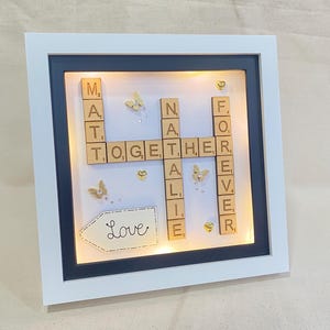 May include: A framed artwork with a white border and a black interior. The artwork features wooden letter tiles spelling "MATT, TOGETHER, ALIE, FOREVER" in a crossword style. Gold butterflies and hearts decorate the piece, with the word "Love" written on a white arrow.