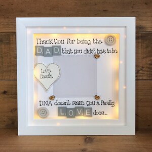 Pu&ograve; includere: Cornice per foto bianca con una luce all'interno. La cornice ha uno sfondo bianco con testo nero che dice "Thank You for being the DAD that you didn't have to be. Love, Camila. DNA doesn't make you a family LOVE does..." Ci sono piccoli cuori di legno e bottoni sparsi intorno al testo.