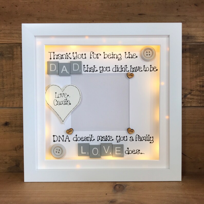 Pu&ograve; includere: Cornice per foto bianca con una luce all'interno. La cornice ha uno sfondo bianco con testo nero che dice "Thank You for being the DAD that you didn't have to be. Love, Camila. DNA doesn't make you a family LOVE does..." Ci sono piccoli cuori di legno e bottoni sparsi intorno al testo.