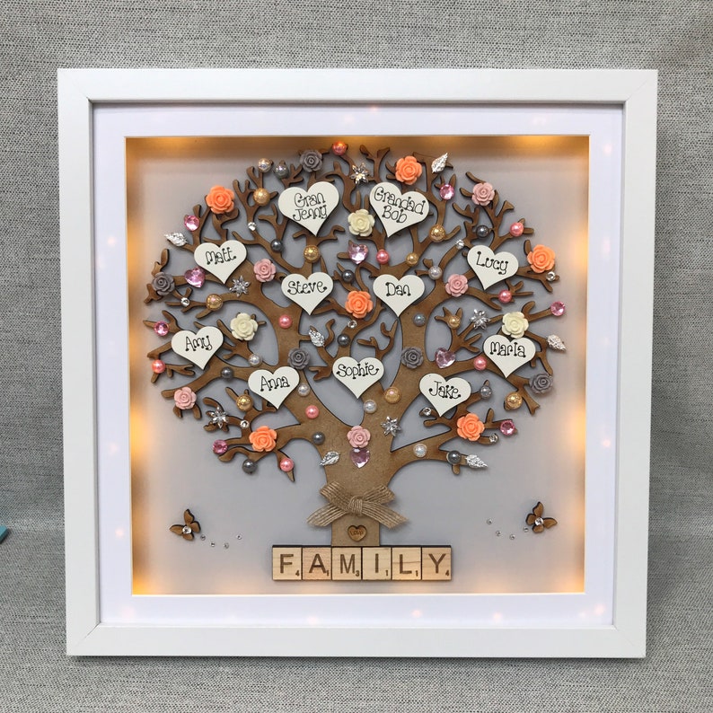 May include: A light-up wooden family tree with names written on hearts and flowers. The tree is decorated with pink, white, and peach flowers, pearls, and glitter. The words "FAMILY." are spelled out in wooden blocks at the bottom of the tree.