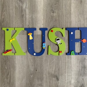 May include: A wooden name sign spelling "KUSH" in green and blue letters with space-themed decorations. The letters are decorated with stars, planets, rockets, and astronauts.