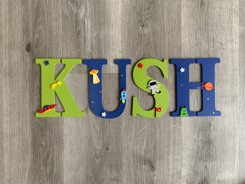 May include: A wooden name sign spelling "KUSH" in green and blue letters with space-themed decorations. The letters are decorated with stars, planets, rockets, and astronauts.