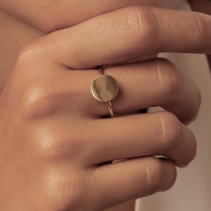 Könnte beinhalten: Ein goldener Ring mit einem flachen, kreisförmigen Scheibenelement auf einer dünnen, polierten Ringschiene. Der Ring wird am Mittelfinger getragen und durch einen passenden goldenen Creolen-Ohrring am Ohrläppchen ergänzt, was ein stimmiges Schmuckset ergibt.