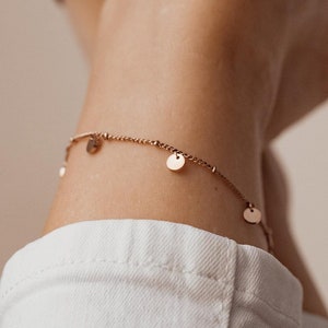 Può includere: Un delicato braccialetto per caviglia in oro con piccoli ciondoli rotondi che pendono dalla catena. Il braccialetto per caviglia è indossato sulla caviglia di una persona.