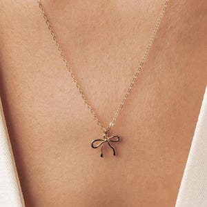 May include: A delicate gold-toned necklace featuring a small, minimalist bow-shaped pendant suspended from a thin, fine-link chain. The metallic finish reflects light, highlighting the simple, symmetrical design of the bow charm centered on the chain.