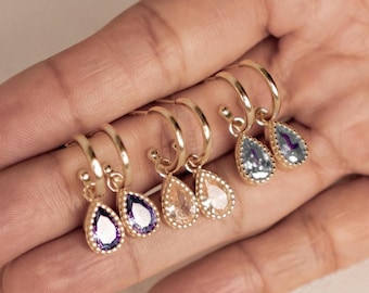 May include: A collection of gold-colored hoop earrings, each adorned with a teardrop-shaped gemstone. The gemstones vary in color, including shades of purple, pink, and blue. The earrings are displayed on a hand against a neutral background.