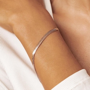 Könnte beinhalten: Ein zartes, dünnes, silberfarbenes Armband mit flachem Schlangenketten-Design. Das Armband wird am Handgelenk getragen. Der Schmuck ist schlicht und elegant, vor weißem Hintergrund.