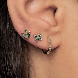 Può includere: Primo piano di un orecchio adornato da tre orecchini. Due sono orecchini a lobo: uno è un design a foglia verde e l'altro è un design a fiore verde. Il terzo è un piccolo orecchino a cerchio dorato con minuscole pietre verdi.