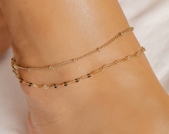 May include: Two delicate gold anklets. The top anklet features small beads, while the bottom one has a chain of flat, elongated shapes. The anklets are worn on a foot, with a soft, white fabric in the background.