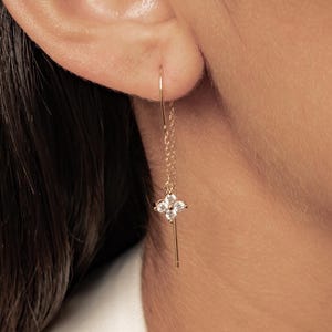May include: A gold threader earring featuring a delicate chain that suspends a small, four-petaled flower charm. The flower is set with four clear, sparkling stones arranged in a clover-like pattern, dangling from a thin, polished gold wire.