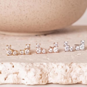 Può includere: Quattro paia di orecchini a lobo con un design a grappolo. Gli orecchini sono dorati e argentati, ciascuno con più pietre chiare e scintillanti. Gli orecchini sono esposti su una superficie chiara e strutturata.