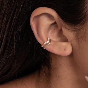 Può includere: Un ear cuff dorato con un design a onda, ornato con piccole pietre preziose trasparenti. Il gioiello è indossato sull'orecchio, mettendo in risalto il suo stile delicato ed elegante. Il cuff avvolge la cartilagine dell'orecchio.