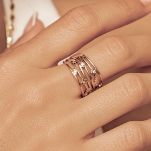 Könnte beinhalten: Ein breiter Goldring mit einem mehrschichtigen, durchbrochenen Design aus mehreren dünnen, miteinander verbundenen Golddrähten. Der Ring ist mit kleinen, runden, klaren Steinen besetzt, die in Abständen entlang der Drahtstruktur sitzen.