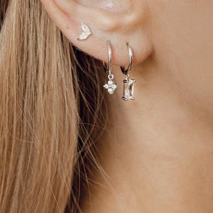 May include: A close-up of a person's ear with three silver earrings. The earrings are a small leaf design, a small diamond-shaped charm, and a larger rectangular charm with a diamond in the centre.