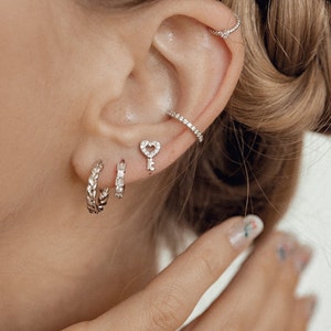 May include: A close-up of a person's ear with multiple silver earrings. The earrings include a small key with a heart-shaped hole, a small hoop, and a larger hoop with a braided design.