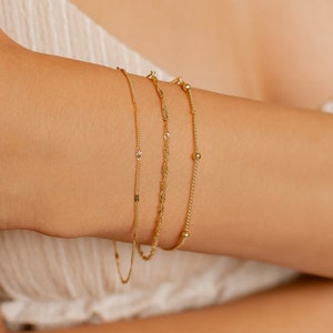 May include: Three gold bracelets stacked on a person's wrist. The bracelets are all different styles, one is a delicate chain, one is a thicker chain with small beads, and the other is a chain with small, square-shaped links.