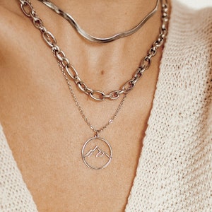 May include: A silver chain necklace with a mountain pendant. The necklace is layered with two other silver chains.