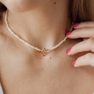 Op de afbeelding: Een delicate parelketting met een gouden sluiting. De ketting wordt gedragen door een persoon met rode nagellak.