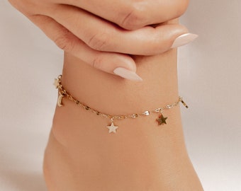 Może przedstawiać: Złota bransoletka na kostkę z delikatnym łańcuszkiem i zawieszkami w kształcie gwiazd. Bransoletka na kostkę znajduje się na kostce osoby.