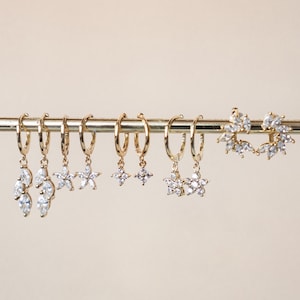 May include: A set of gold hoop earrings with dangling charms. The charms include a cluster of pear-shaped stones, a star, and a flower.