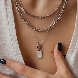 Könnte beinhalten: Eine silberne Kettenkette mit einem mehrschichtigen Design. Die Kette hat einen Anhänger mit einem rechteckigen weißen Stein.