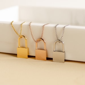 May include: Three padlock pendant necklaces in gold, rose gold, and silver. Each necklace features a small, rectangular padlock pendant suspended from a delicate chain. The pendants are displayed on a white surface against a soft, neutral background.