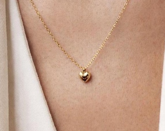May include: A delicate gold necklace featuring a small heart pendant. The pendant is centered on a fine chain, creating a minimalist and elegant design. The necklace is worn against a white blazer.