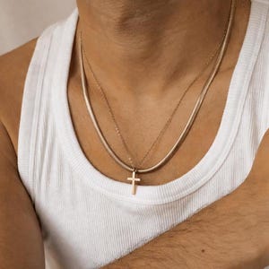 Pode incluir: Dois colares dourados. O colar superior é uma fina corrente de cobra. O colar inferior tem um pequeno pingente de cruz. A pessoa está usando uma camiseta regata branca.