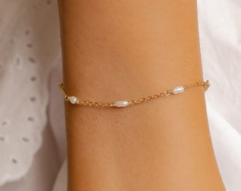 Puede incluir: Un delicado brazalete de cadena dorada con pequeños acentos de perlas blancas. El brazalete se muestra en la muñeca de una persona.