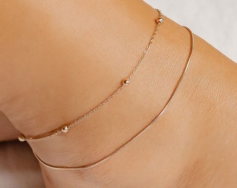 May include: Two gold anklets on a person's ankle. The top anklet has small gold beads and the bottom anklet is a thin chain.