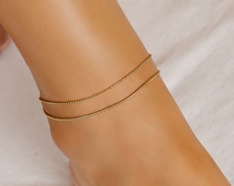 May include: Two gold anklets on a person's ankle. The anklets are layered, one is a thin chain and the other is a thicker chain.