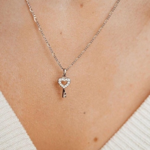 Könnte beinhalten: Silberfarbene Halskette mit einem Schlüsselanhänger. Der Schlüsselkopf ist herzförmig und mit kleinen, klaren Edelsteinen besetzt. Die Kette wird vor einem weißen Pullover getragen.