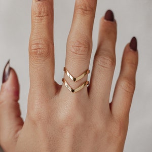 Puede incluir: Anillo de doble chevron dorado en un dedo. El anillo está hecho de dos bandas finas que forman una forma de chevron.