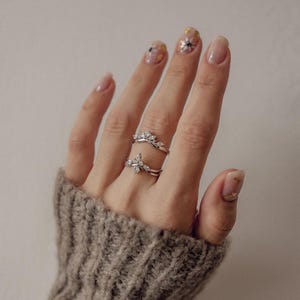 May include: Two silver rings with clear gemstones are displayed on a hand wearing a gray sweater. The rings feature a floral design. The nails are painted with a light pink polish and gold accents.