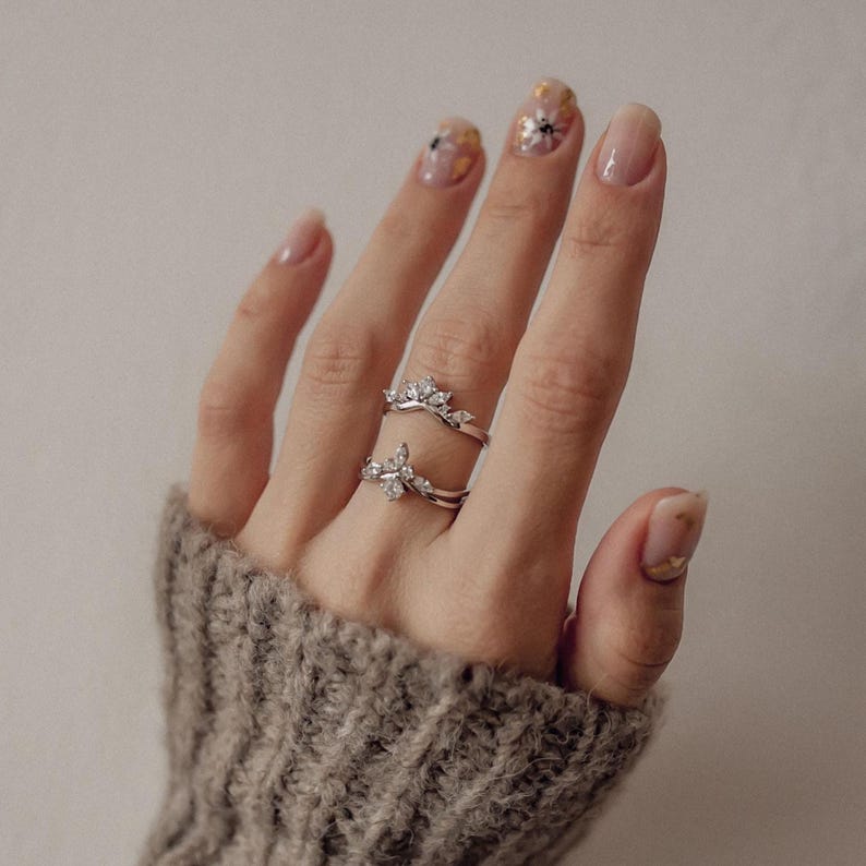 May include: Two silver rings with clear gemstones are displayed on a hand wearing a gray sweater. The rings feature a floral design. The nails are painted with a light pink polish and gold accents.