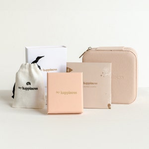 May include: A pink and white gift box set with the text "hey happiness" on the front of each box. The set includes a small pouch, a jewelry box, and a polishing cloth.