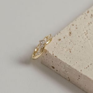 May include: A gold-colored ring with a floral design, featuring multiple clear, sparkling gemstones. The ring is set against a neutral, textured background, highlighting its intricate details and elegant style.