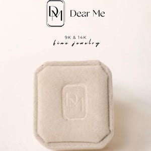 K&ouml;nnte beinhalten: Eine quadratische Ringbox aus wei&szlig;em Samt mit abgerundeten Ecken. Die Box hat die Initialen "DM" auf der Oberseite gepr&auml;gt. Der Text "Dear Me 9K & 14K fine jewelry" ist &uuml;ber der Box gedruckt.