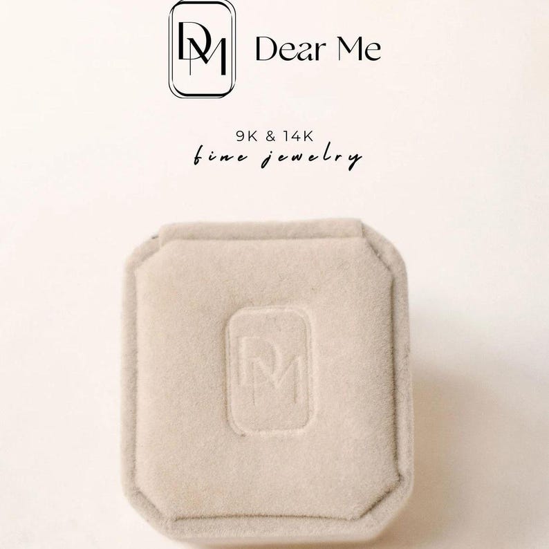 Op de afbeelding: Een vierkante ringdoos van wit fluweel met afgeronde hoeken. De doos heeft de initialen "DM" in reli&euml;f op de bovenkant. De tekst "Dear Me 9K & 14K fine jewelry" is boven de doos gedrukt.