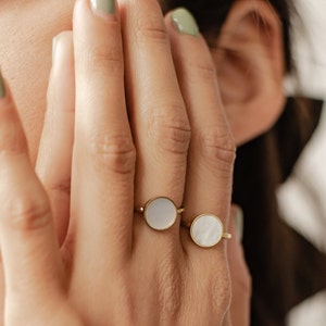 K&ouml;nnte beinhalten: Zwei goldene Ringe mit wei&szlig;en runden Steinen. Die Ringe befinden sich an einem Finger.