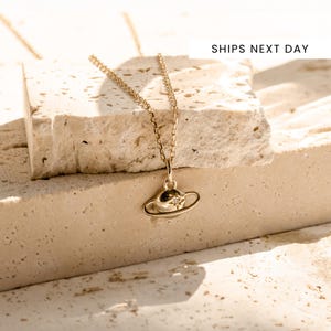 Puede incluir: Un collar dorado con un colgante de planeta. El colgante presenta un diseño de planeta con un anillo y una pequeña gema brillante. El collar se muestra sobre una superficie texturizada y de color claro.